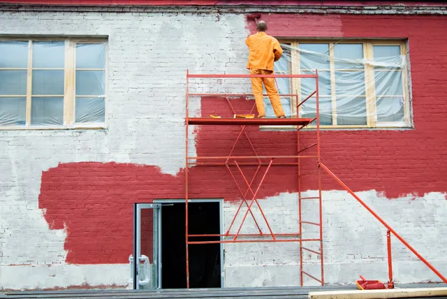 Peintre sur échafaudage appliquant une peinture rouge sur la façade en briques d’un bâtiment